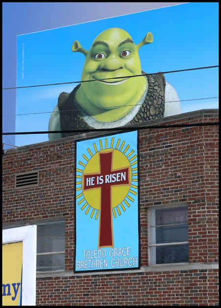 Photo: Shrek billboard above a a church banner reading 'He Is Risen'.