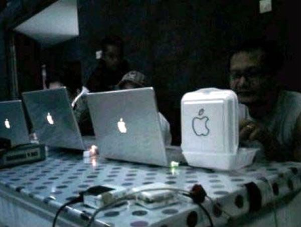 Three people working on Powerbooks with the Apple logo glowing on their cover, and one guy eating lunch from a styrofoam container with the Apple logo drawn on it in magic marker.