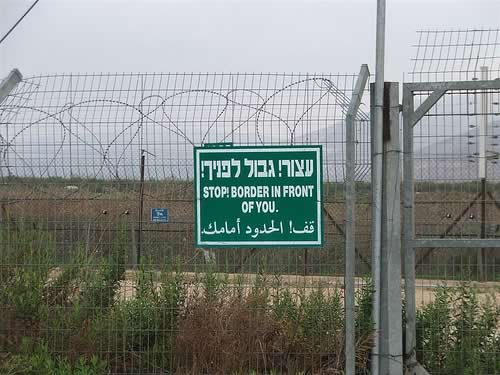 Photo of chain link/barbed-wire fence along Israel/Lebanon border with a sign that reads 'Stop! Border right in front of you'.