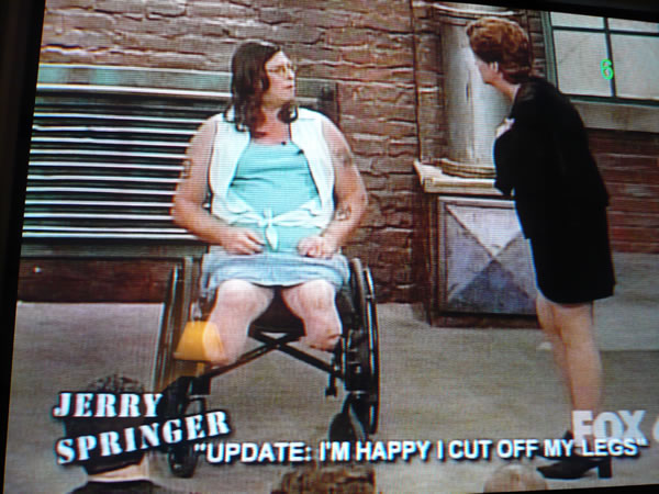 Scene from “Jerry Springer” with man in woman’s clothing, sitting in a wheelchair with severed legs.