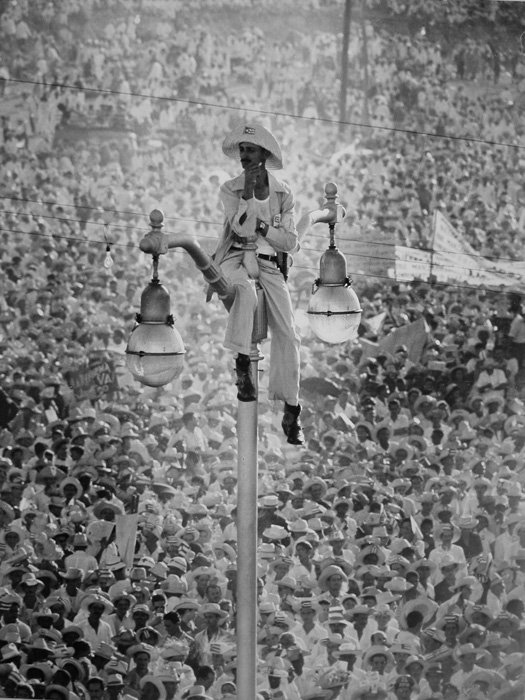 Man in white clothes and white hat, sitting atop a lamp post over a crowd in white clothes and white hats.