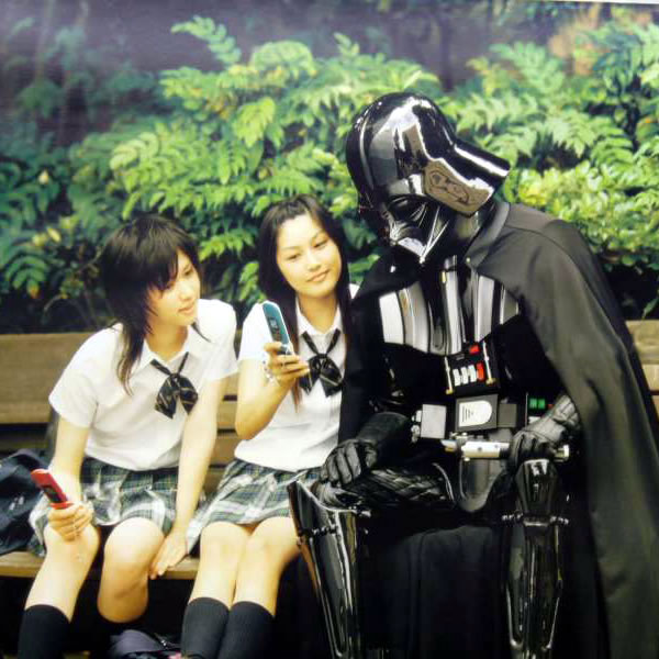 Two Japanese schoolgirls, sitting on a park bench with Darth Vader, showing him pictures on their cellphones.