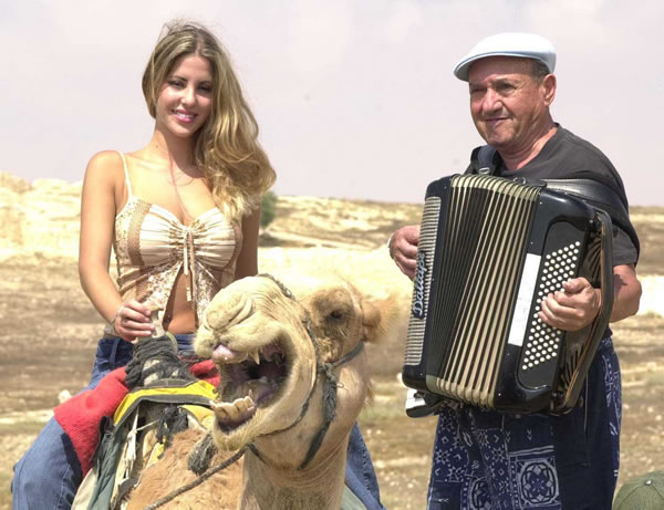 Older man playing accordion beside a smiling young blonde woman on a camel