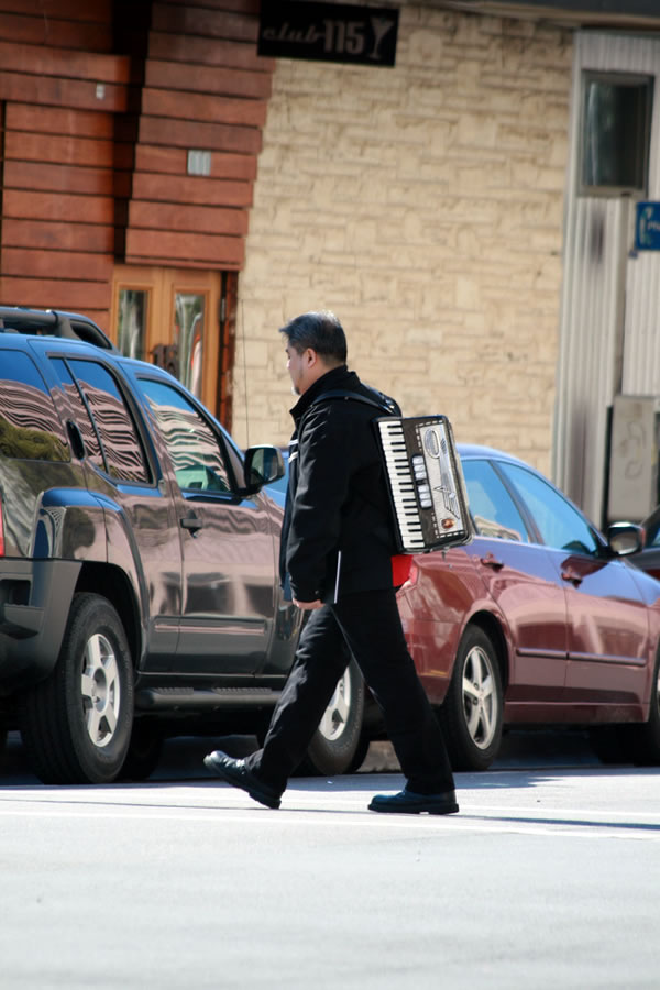 Joey deVilla, dressed all in black, walking down an Austin street with an accordion on his back.