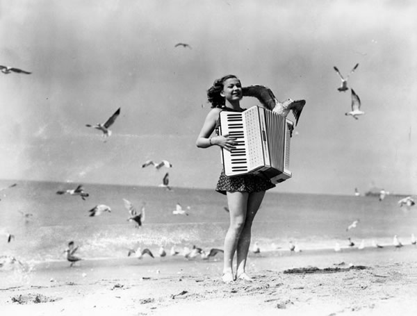 Old black-and-white photo of a woman playing accordion on a beach.