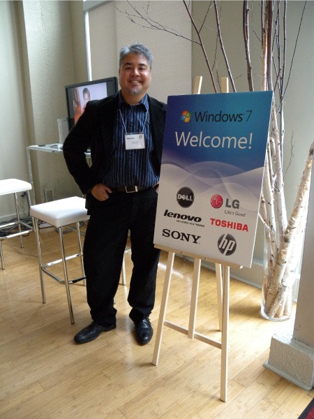 Joey devilla, standing beside a "Windows 7: Welcome!" sign