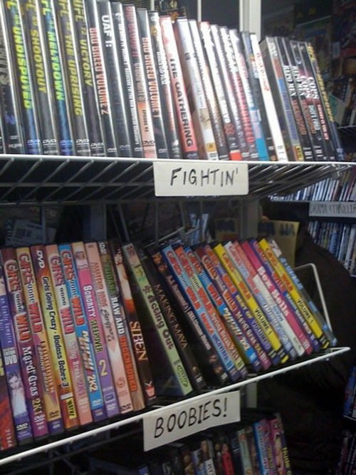Two DVD shelves at a video store, one labelled 'Fightin', the other labelled 'Boobies!'