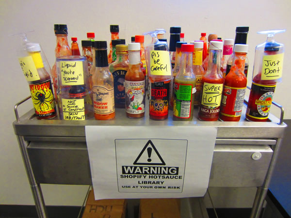 Tabletop packed with various bottles of hot sauce. A sign below it reads "Warning: Shopify hotsauce library. Use at your own risk."