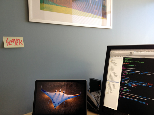 Photo of an office scene with computer monitors, bland framed office art and a post-it note with the "Slayer" logo drawn on it in red marker