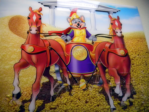 Portrait of Chuck E. Cheese dressed up as a Roman gladiator riding a chariot drawn by two horses. The road on which he rides is made of Chuck E. Cheese game tokens.