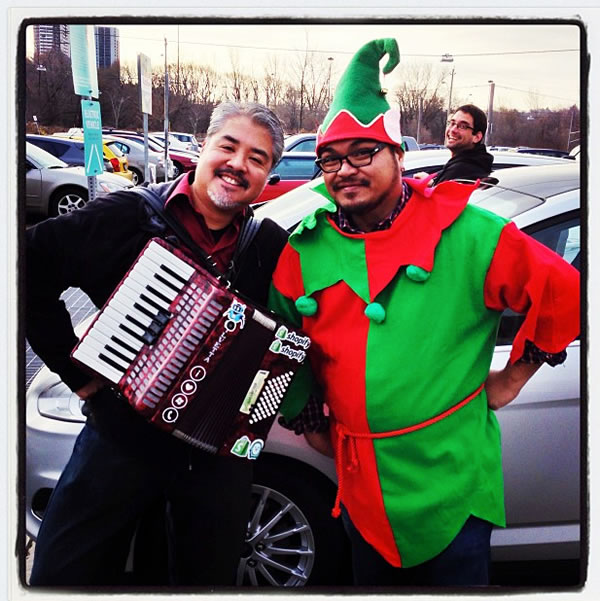 Joey deVilla with his accordion, posing with Rannie Turingan dressed as an elf. In the background, Sean Carruthers looks on.