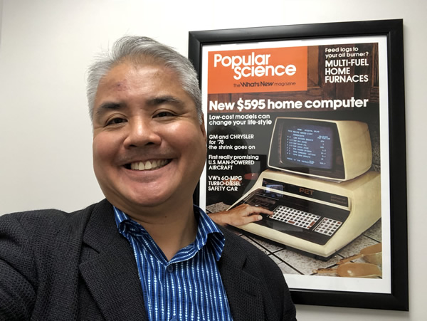 Joey deVilla smiles beside a framed poster featuring the 1977 issue of Popular Science featuring the Commodore PET 2001 computer.