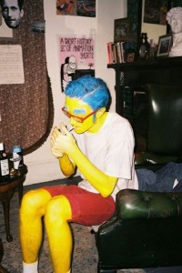 Guy dressed up as "Milhouse" from "The Simpsons", in full yellow body paint, with blue hair and blue exaggerated eyebrows, wearing a white t-shirt and shorts, sitting down an lighting a cigarette.