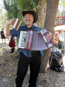 Joey deVilla in a "Three Musketeers"-style hat with feathers, holding a wooden mug with a carved dragon handle, playing the accordion and smiling