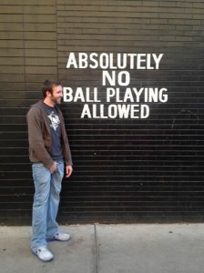 Smiling guy with his hands down his pants in front of a wall with "Absolutely no ball playing allowed" painted on it.