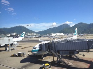 Tarmac at Hong Kong airport, with parked planes in the foreground and mountains and a plane taking off in the background