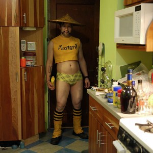 Asian man in Chinese farmer's hat, yellow crop-top T-shirt that says "Mustard", too-short yellow shorts and yellow socks, posing in the kitchen with a bottle of mustard, all in a very disturbing fashion.