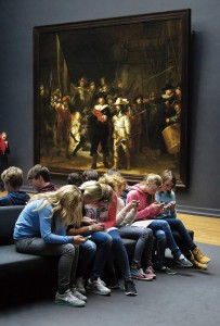 Photo: Schoolchildren sitting on a bench in a museum, engrossed in their smartphones, completely ignoring the large masterpiece hanging on the wall in the background.