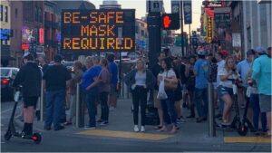 Photo: Street scene in US city. Large highway sign reads “Be safe - Mask required”, but nobody is the picture is wearing a mask.