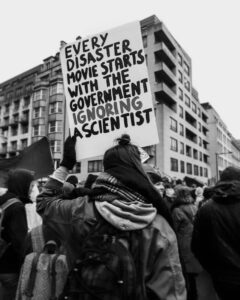 Photo: Protestor holding up sign that reads “Every disaster movie starts with the government ignoring a scientist”.