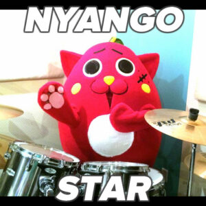 Photo: “Nyango Star” — a costumed mascot that looks like a cat/apple hybrid, sitting at a drum kit.