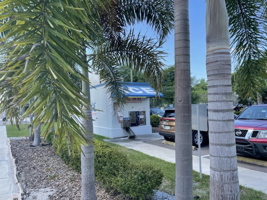 The automated ice dispensing station on Florida Avenue, as seen from the sidewalk through some palm trees.