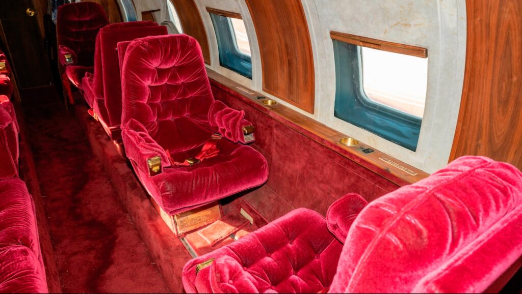 Angle view from the aisle showing two face-to-face red velvet seats. There is a lot of wood paneling.