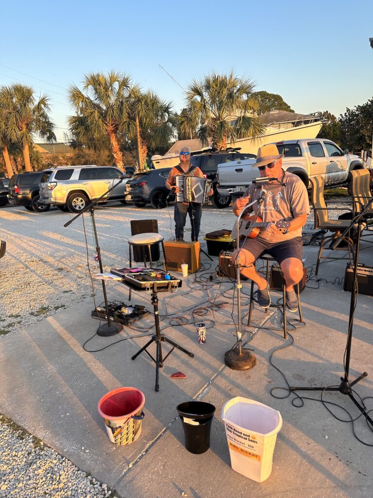 Tom Hood plays ukulele in the foreground as Joey deVilla plays accordion in the background. Behind them is a gravel parking lot with cars, a boat, and palm trees.