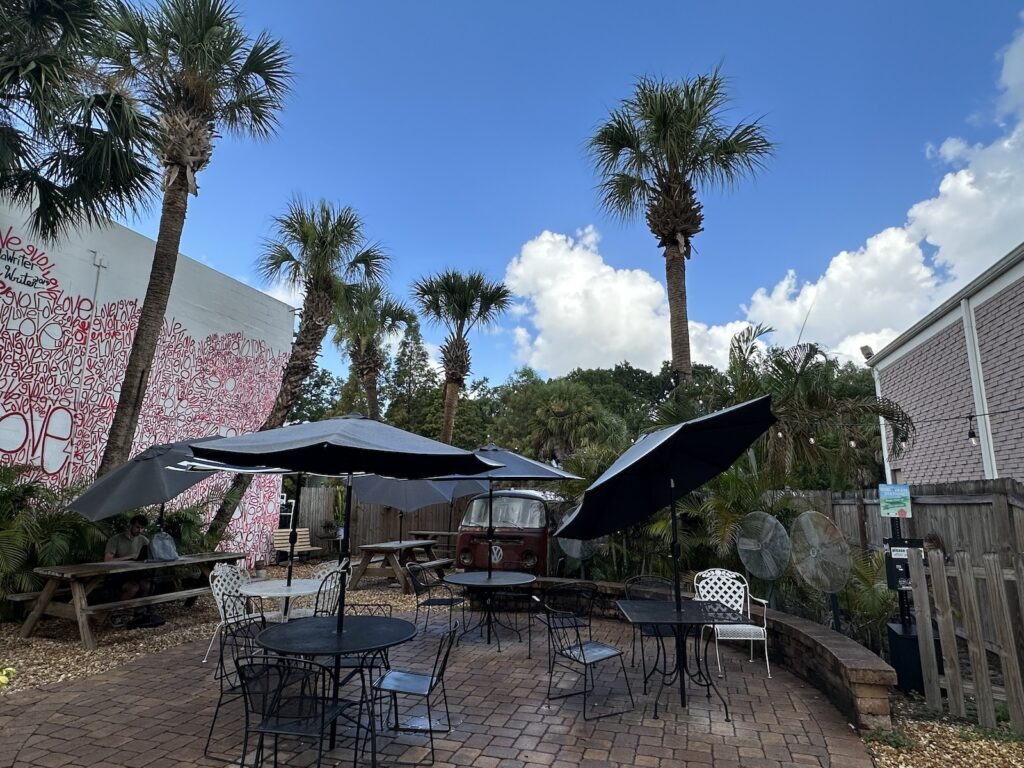 The Spaddy’s Coffee courtyard, an open area tiled with brick featuring outdoor tables and chairs with umbrellas, and palm trees in the background.