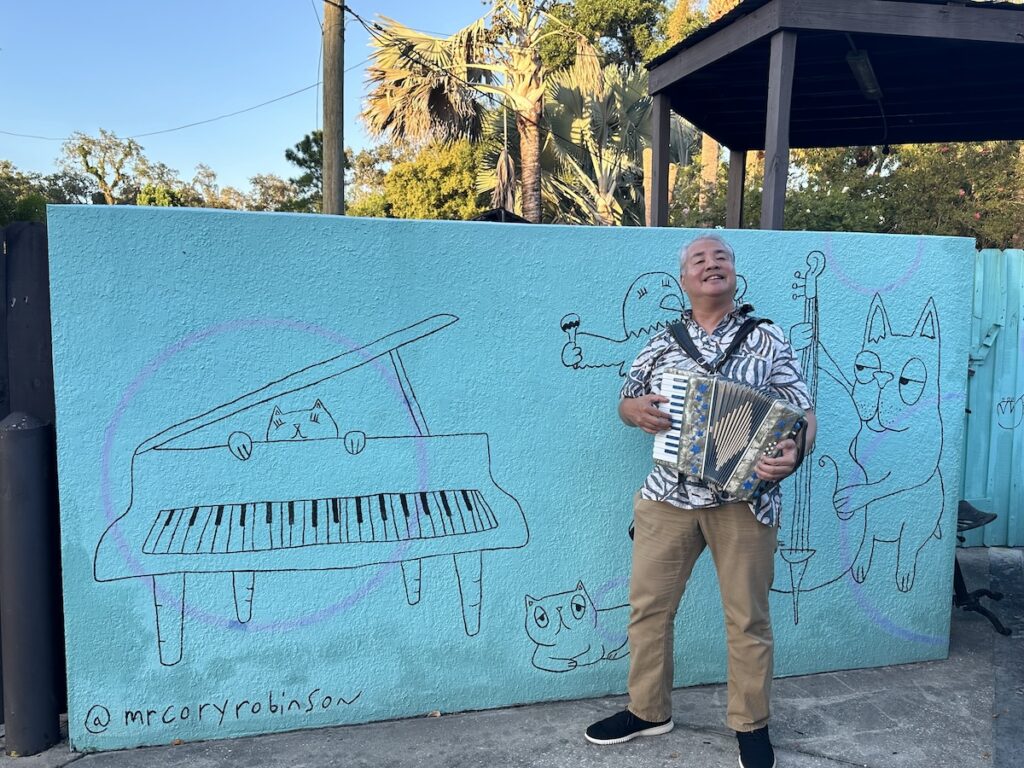 Joey de Villa playing accordion beside the blue wall with cartoon animal musicians behind Ella’s Americana Folk Art Cafe.